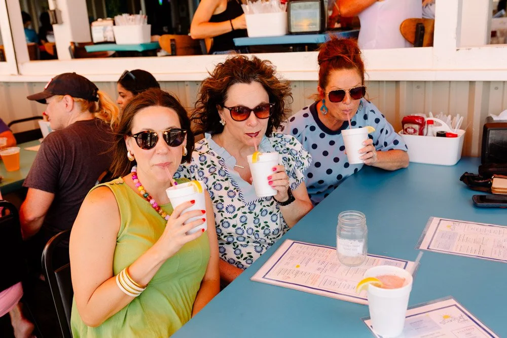 Three women sitting at a blue table enjoying drinks, with a woman in the background at a counter. The women are wearing sunglasses and sipping drinks through straws, with menus on the table.