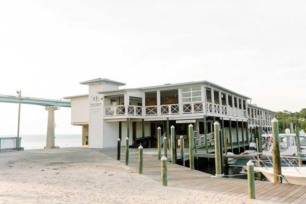 A two-story seaside restaurant and bar with white wood siding and a wraparound porch, docks with boats in the marina, and a bridge in the background.