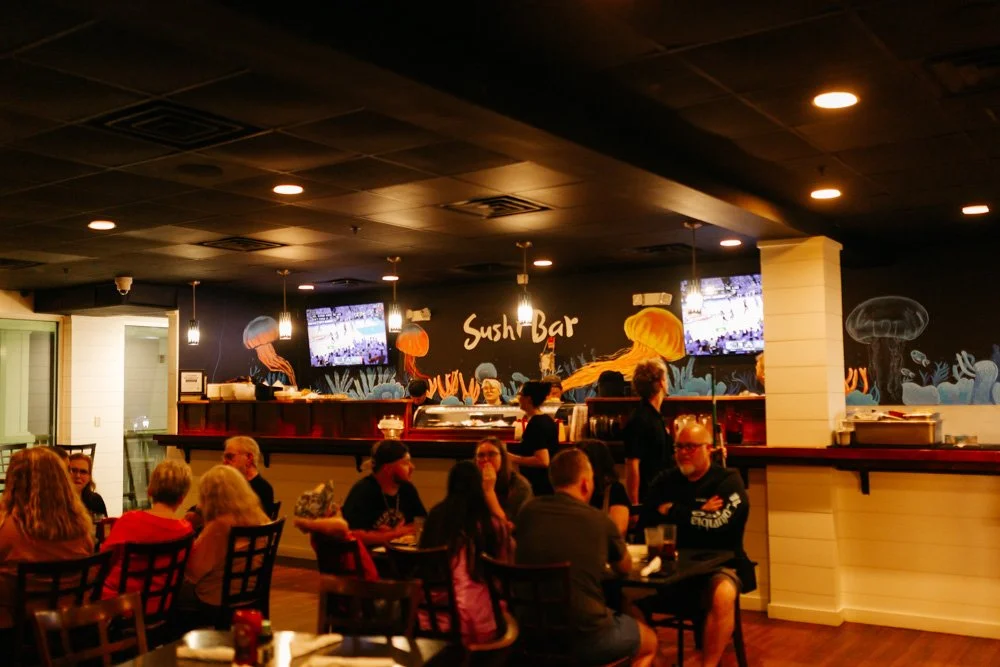 Inside a sushi bar with a black wall decorated with colorful jellyfish murals, patrons dining at dark wood tables, and TV screens hanging above the bar area.
