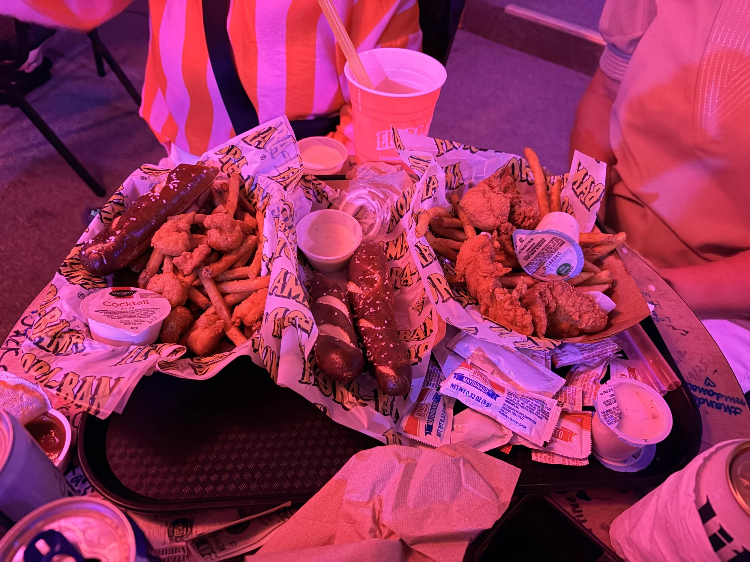 A tray of fast food with fried chicken tenders, sausage, a hoagie sandwich, and condiments, on a table at a restaurant.