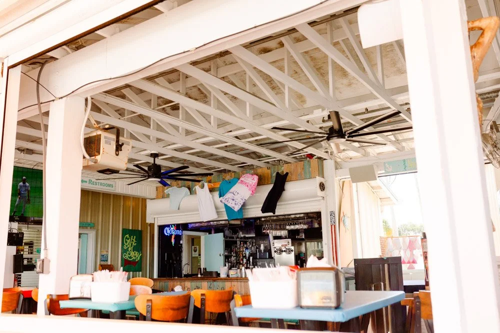Interior of a bar or restaurant with wooden chairs and tables, ceiling fans, and hanging surfboards or boogie boards.