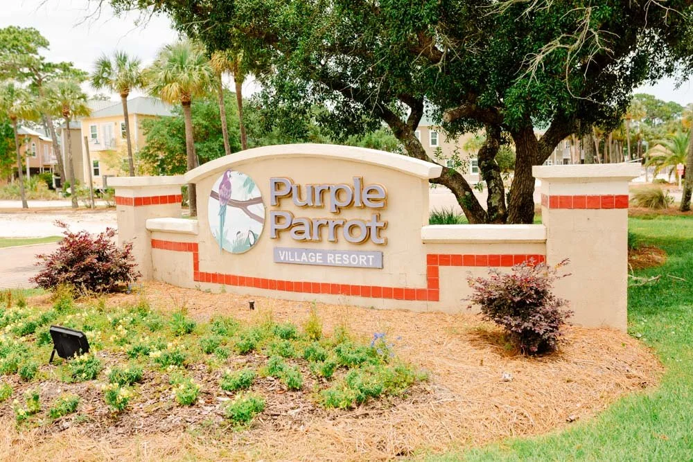 Entrance sign for Purple Parrot Village Resort, set in landscaped area with bushes, trees, and a background of buildings and palm trees.