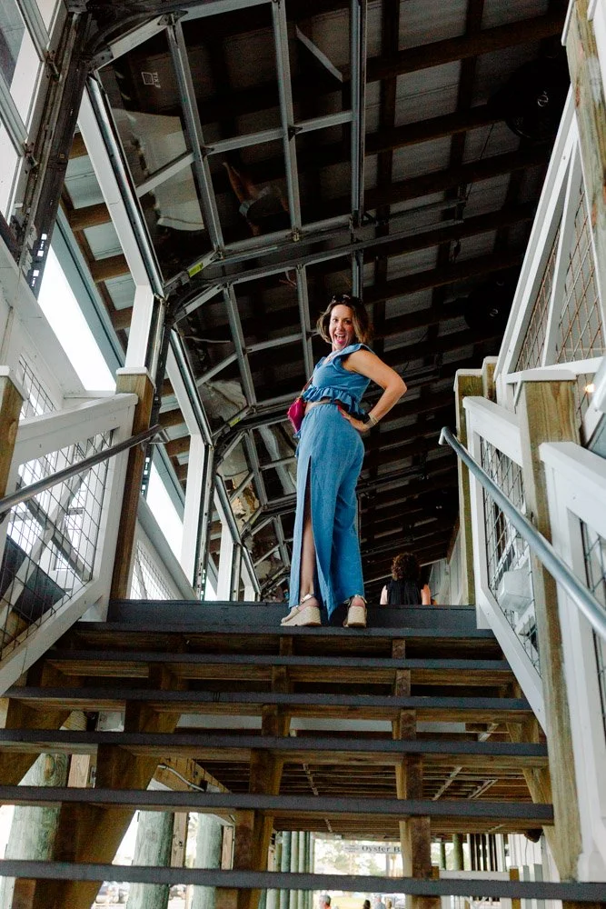Woman standing on a staircase under a covered outdoor area, smiling and looking at the camera, with a person in the background facing away.