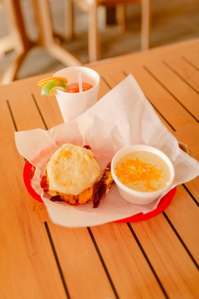 A meal on a wooden table, including a biscuit with gravy, a bowl of yellow rice, and a drink with lime and lemon slices.