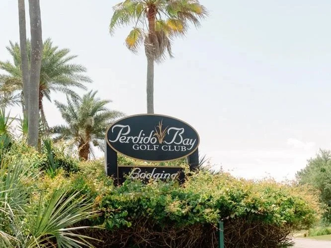 Sign for Perdido Bay Golf Club surrounded by lush greenery and palm trees.