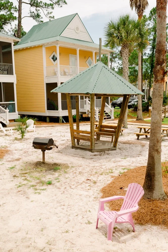 A tropical outdoor setting featuring a yellow beach house with white accents, a small wooden gazebo with a green metal roof, pink Adirondack chairs, a barbecue grill, and tall trees with lush foliage.