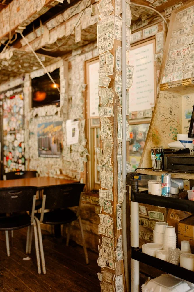 Interior of a rustic cafe or restaurant with a wall decorated with dollar bills, a table with chairs, and a counter with cups and supplies.