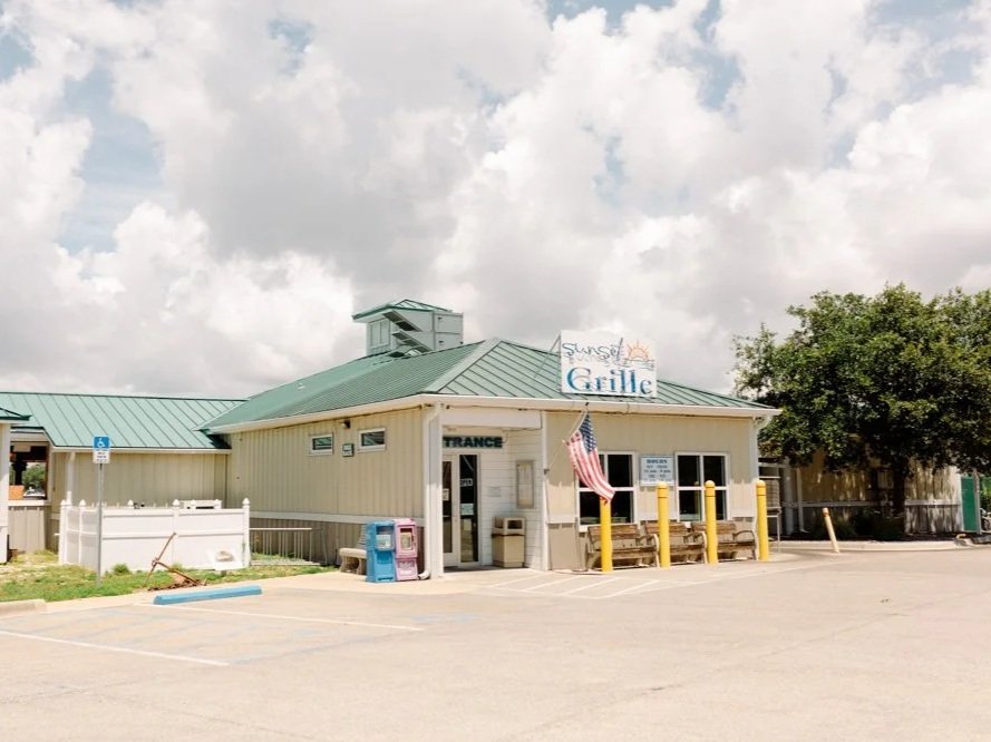 A small restaurant named Sunset Grille with a green metal roof and beige walls, located in a parking lot under partly cloudy skies. The entrance has a sign reading "TRANCE," and an American flag is displayed near the front. There are yellow bollards, a bench, and trash cans outside.