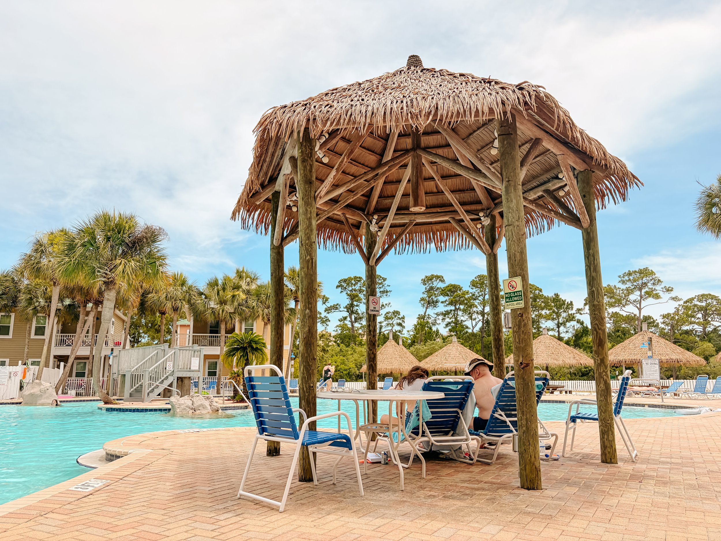A swimming pool area with lounge chairs and a thatched roof tiki hut providing shade. Several people are relaxing by the pool, and there are palm trees and buildings in the background.