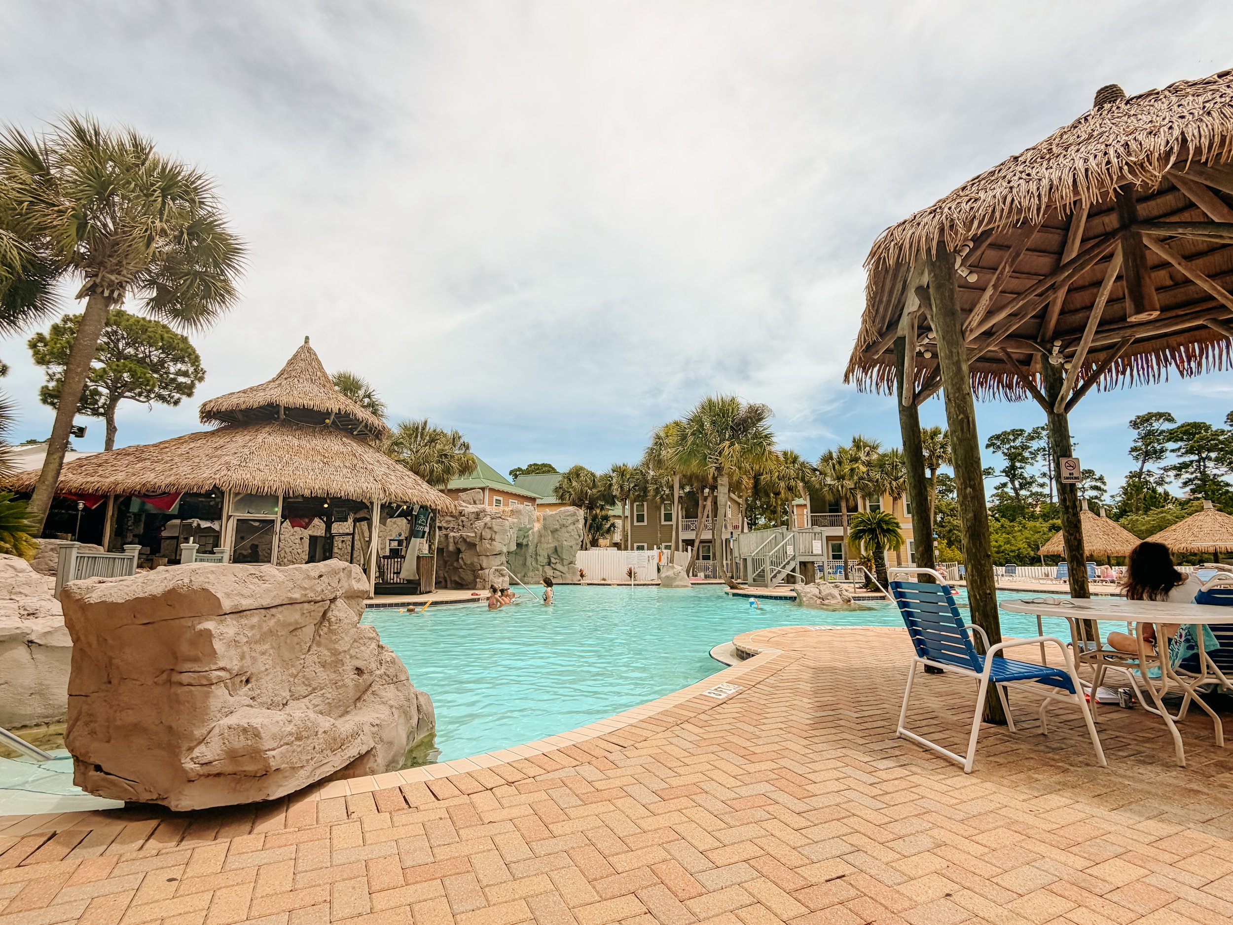 A swimming pool area with thatched-roof huts, lounge chairs, and palm trees under a partly cloudy sky.
