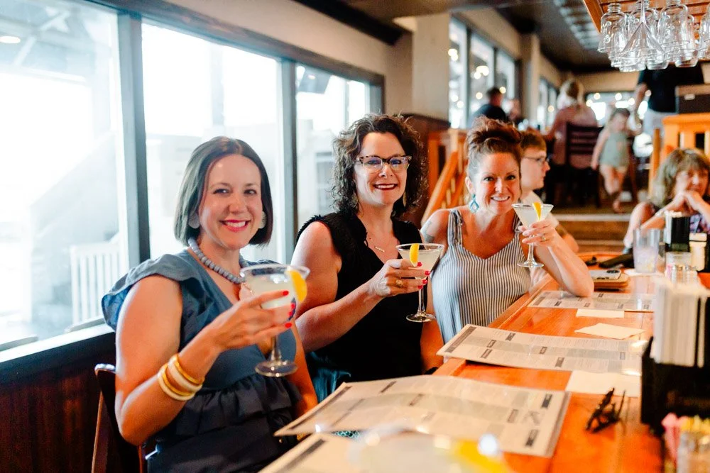 Three women sitting at a restaurant bar holding cocktails and smiling.