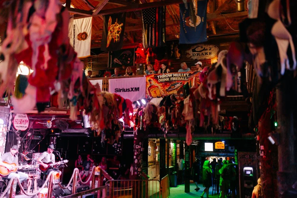 Interior of a bar or music venue with hanging fabric decorations, people watching a live band on stage, and flags hanging from the ceiling.