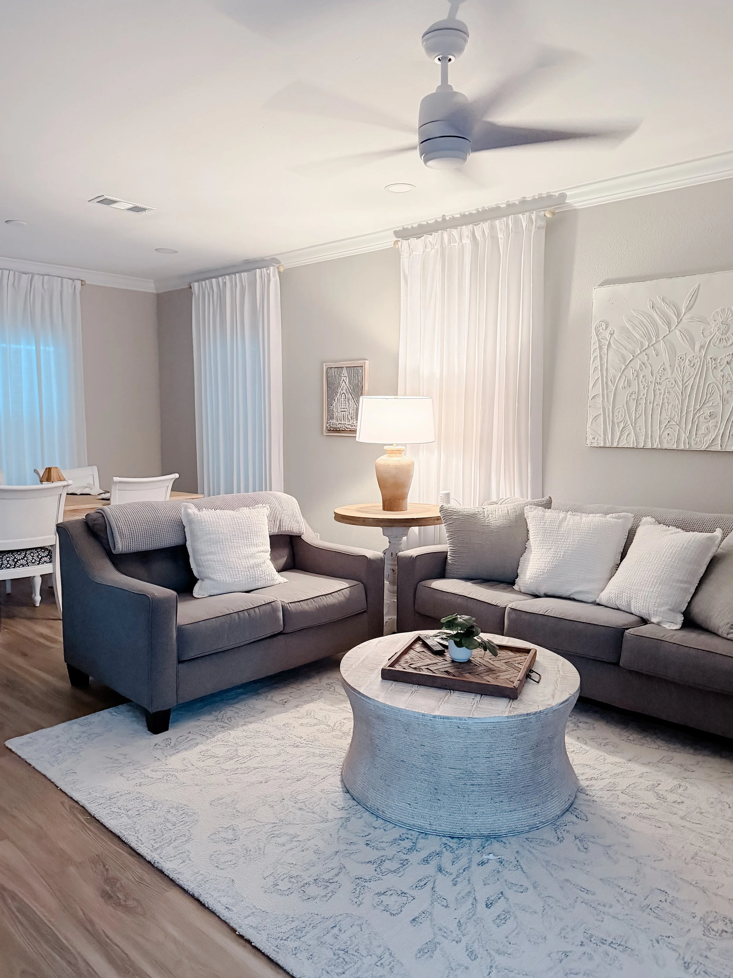 Living room with gray sofas, white pillows, a round coffee table with a tray and a plant, a table lamp, white curtains, wall art, and a ceiling fan.
