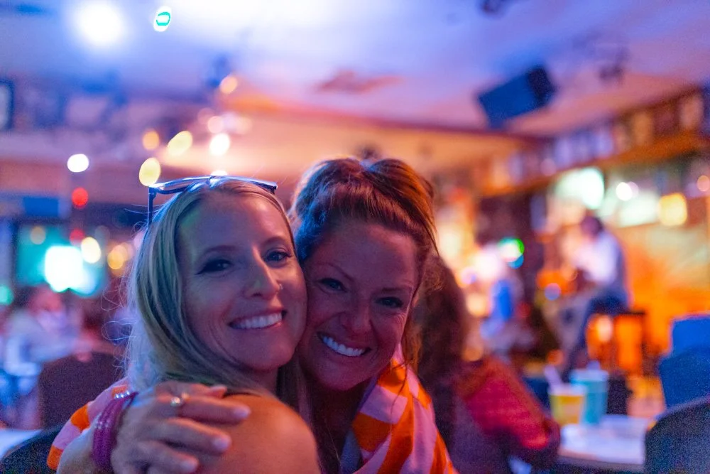 Two women smiling and hugging in a lively bar or restaurant with colorful blurred background.