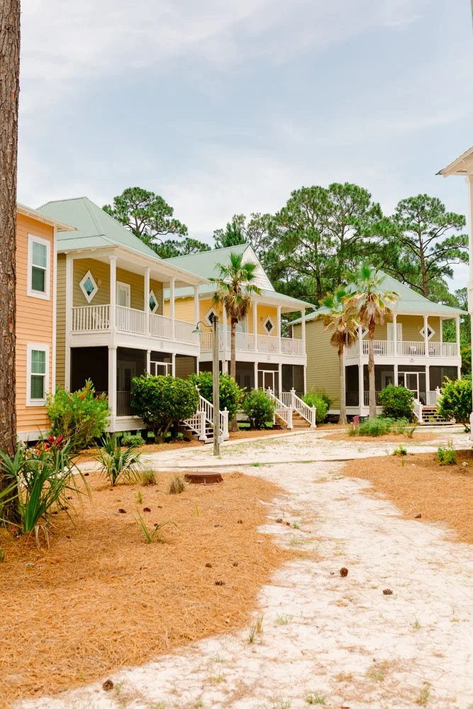 Beachside houses with pastel-colored exteriors, balconies, and palm trees on a sandy pathway under a cloudy sky.