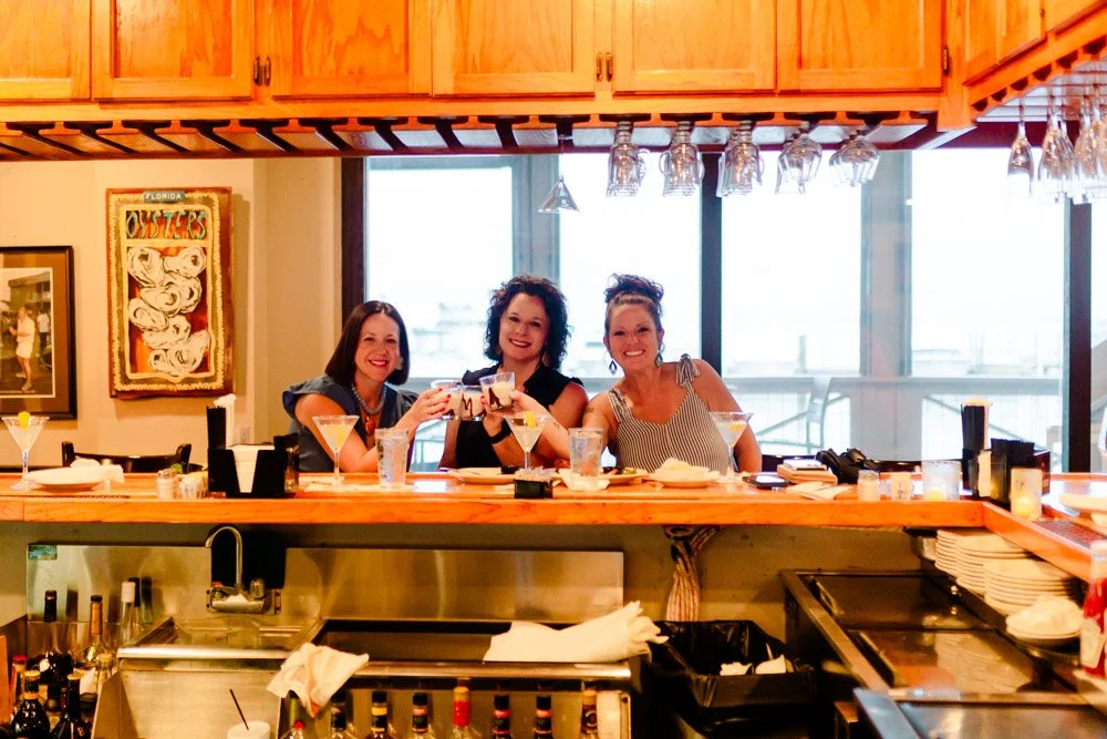 Three women sitting behind a bar with drinks, smiling and raising glasses, in a restaurant with wood cabinetry, large windows, hanging glasses, and art on the wall.