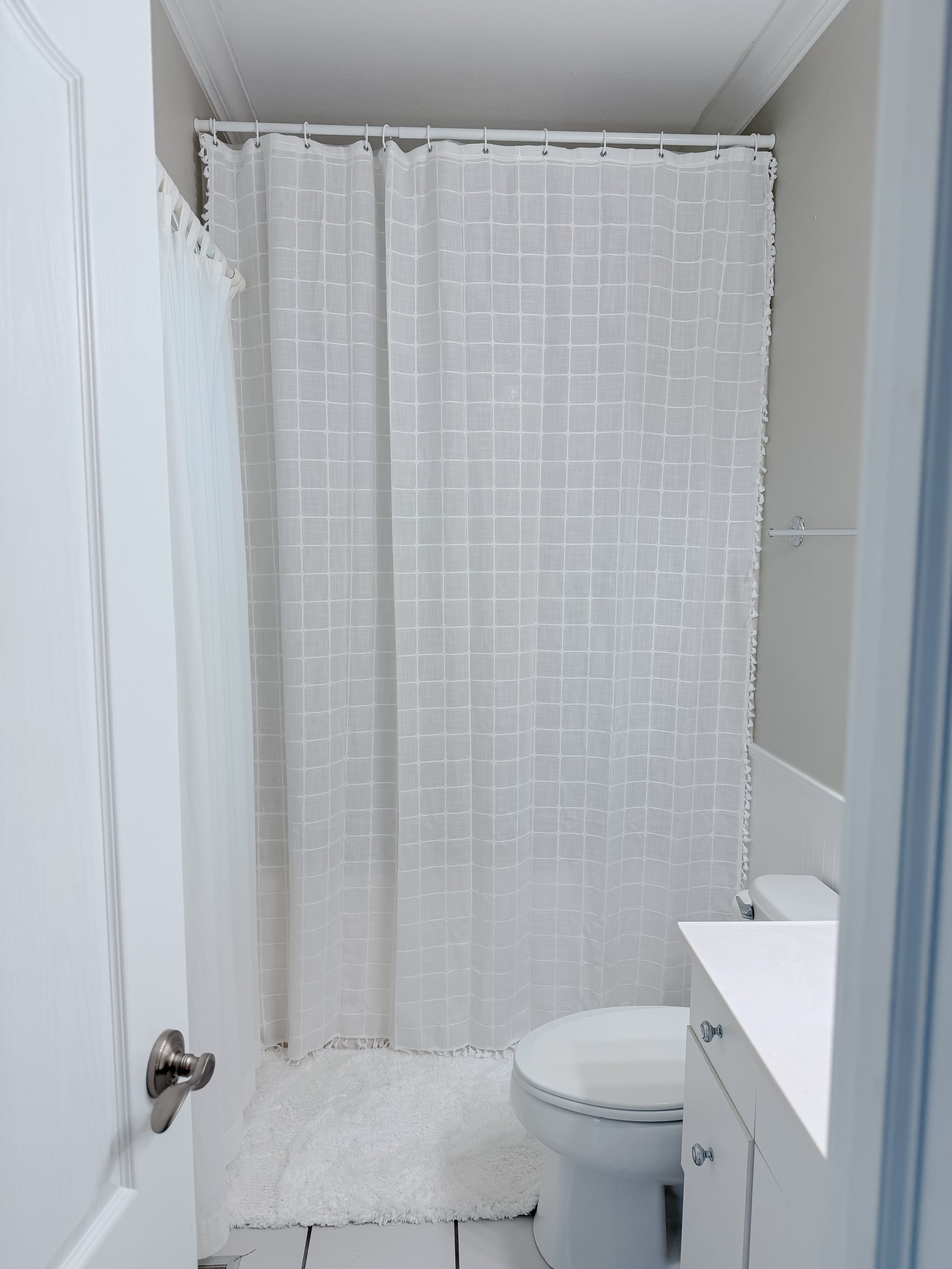 Bathroom with a white shower curtain, white toilet, white vanity with drawers, white fluffy rug, and gray walls.