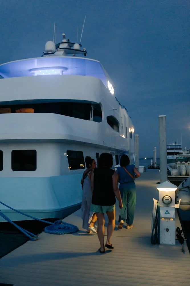 People walking on a marina dock towards a large white yacht during dusk or evening.