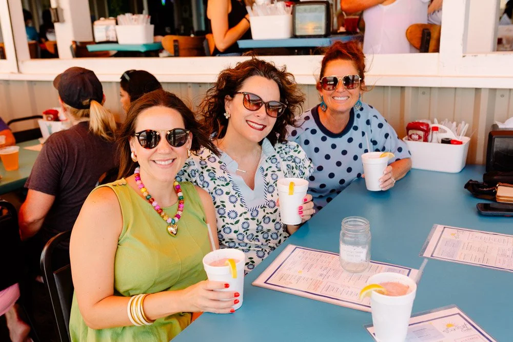 Three women sitting at a blue table in a restaurant, smiling and holding drinks, wearing sunglasses and colorful clothing.