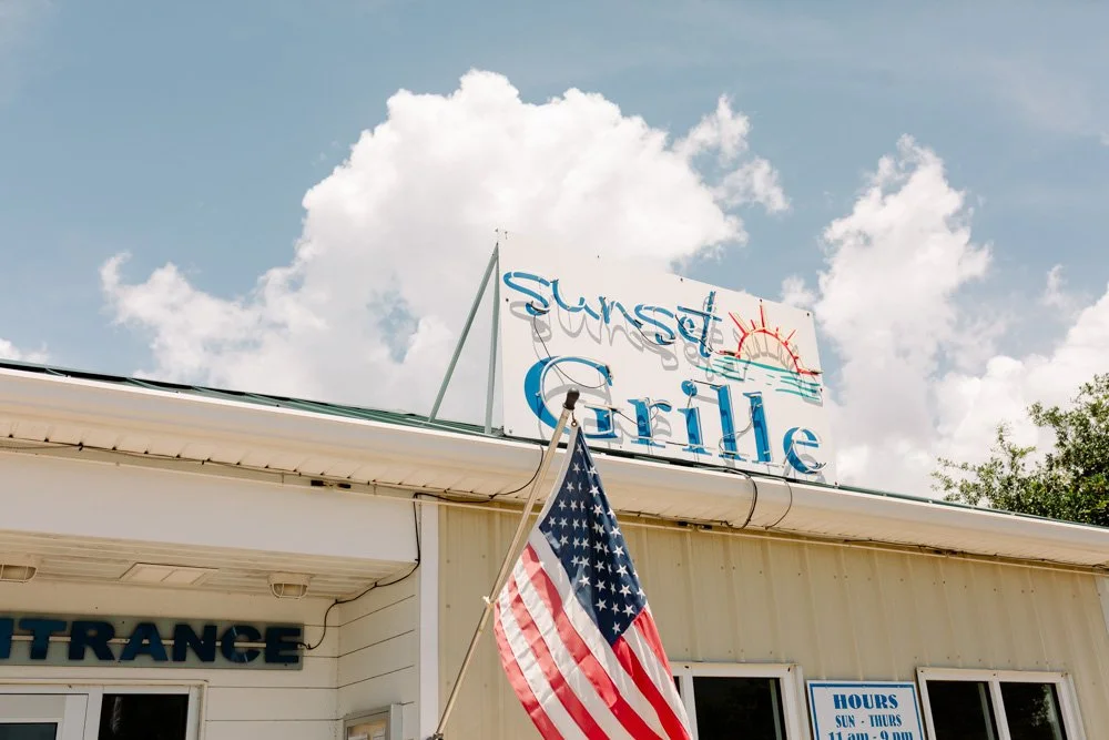 Sunset Grill restaurant sign with a rising sun graphic, American flag in front of the building, and hours posted on the window.