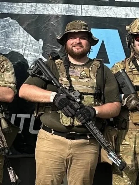 A man wearing tactical gear, a camouflage hat, and gloves, holding a rifle, standing between two other individuals in military-style uniforms.