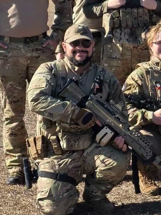 A group of soldiers in camouflage uniforms, one kneeling with a large firearm, outdoors on a sunny day.