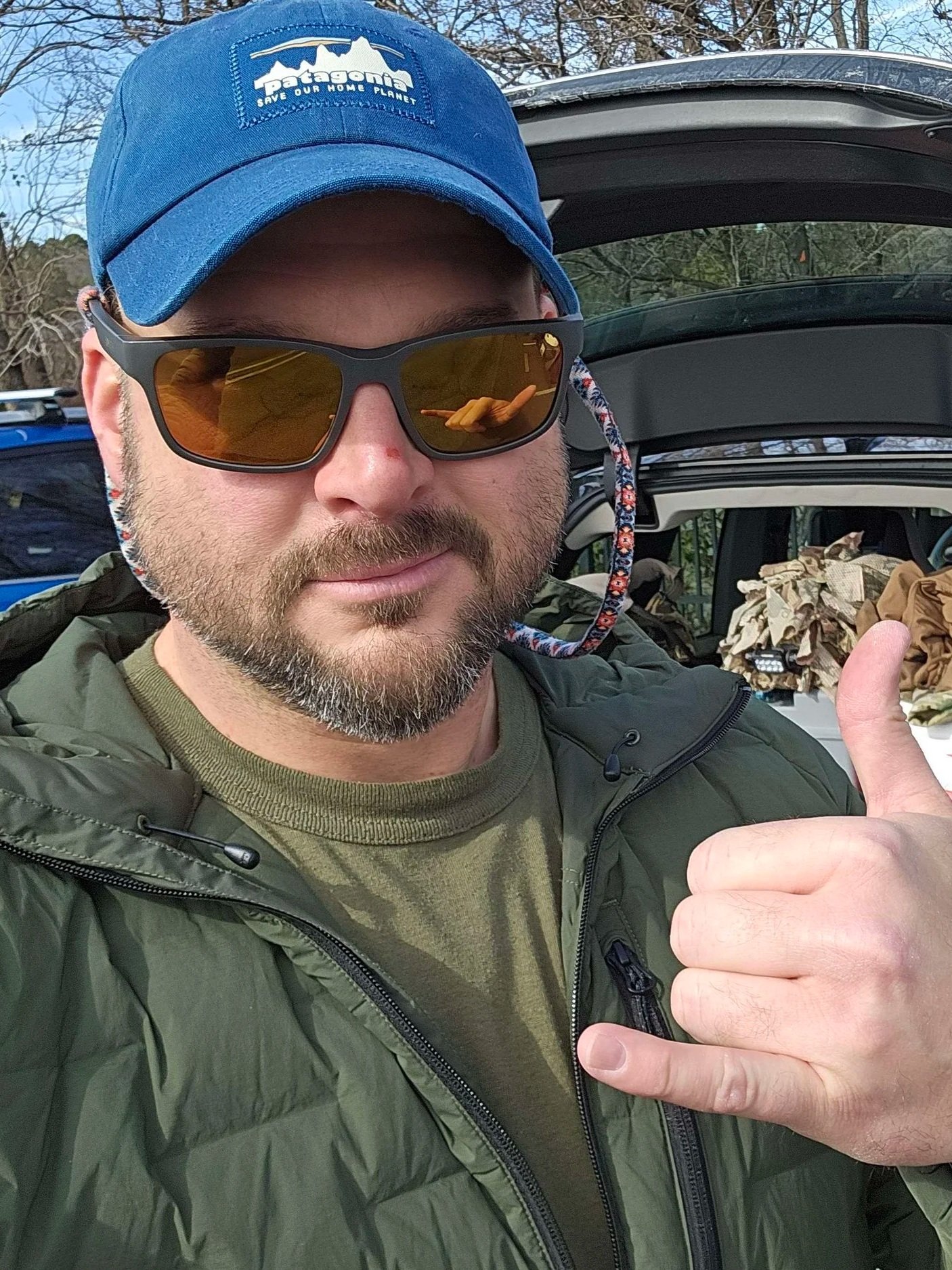 A man wearing a blue Patagonia cap, sunglasses with a reflection of his hand, a green jacket, and an olive green shirt giving a thumbs-up gesture. He is standing outdoors with trees and a car trunk in the background.