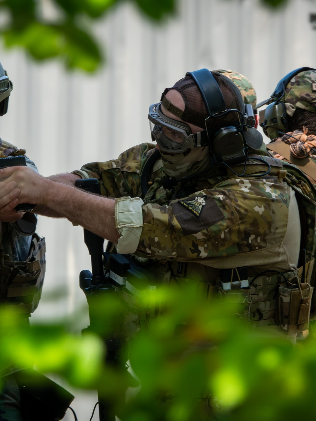 Airsoft player in camouflage uniform wearing tactical gear and communication equipment, exchanging a handshake with another soldier.