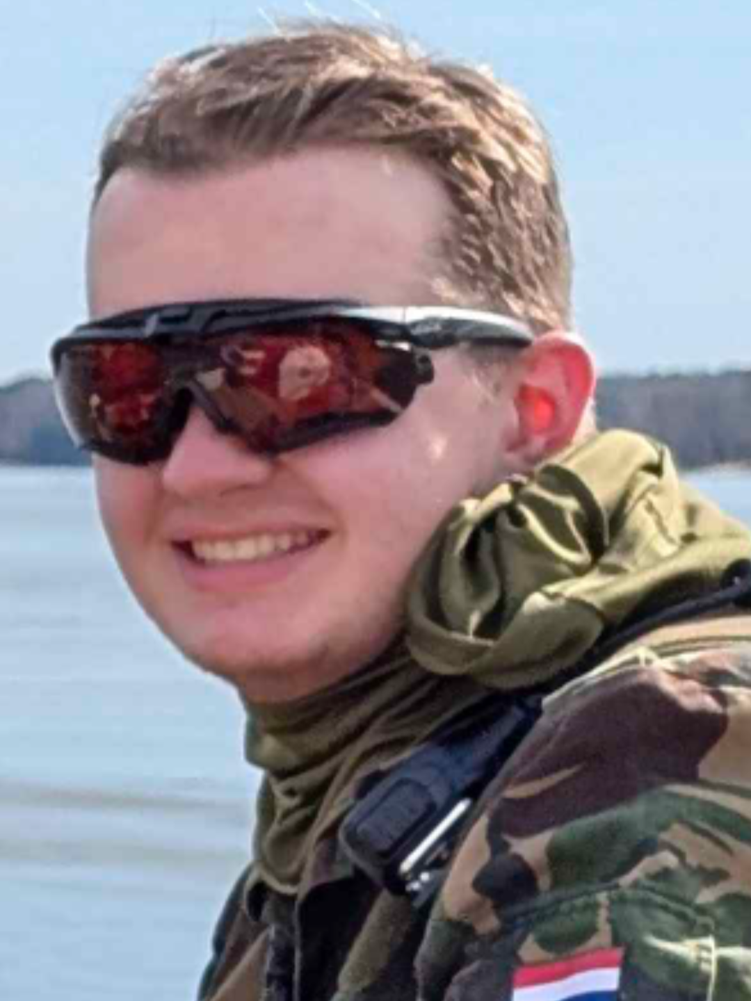 A person smiling wearing sunglasses with a water body in the background.