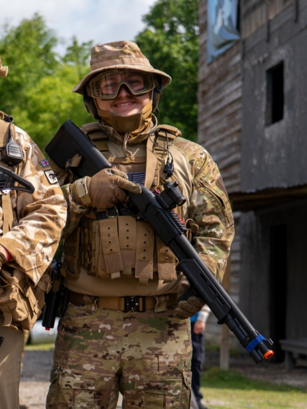 Airsoft player in camouflage uniform and helmet holding a black rifle, standing outdoors.