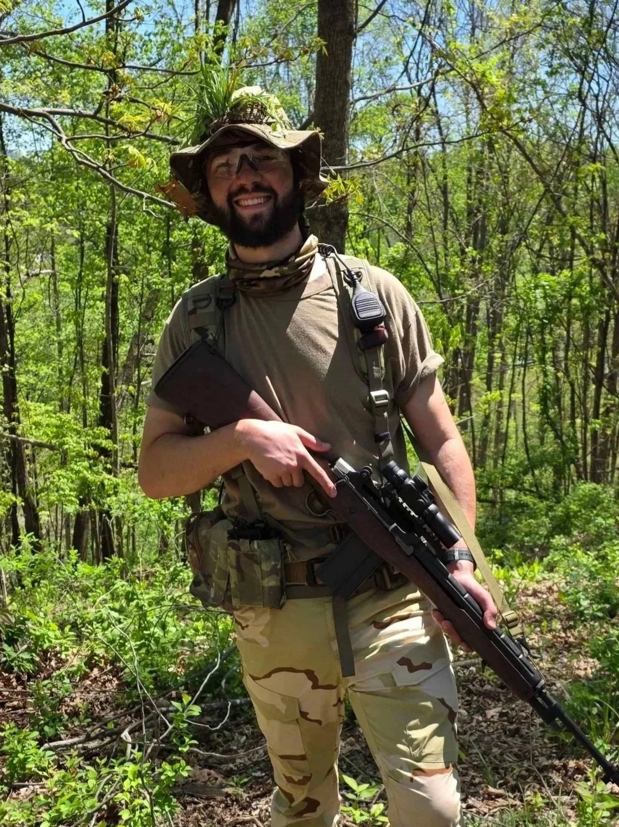 A man dressed in military camouflage gear holding a rifle, standing in a forest with green trees and foliage.