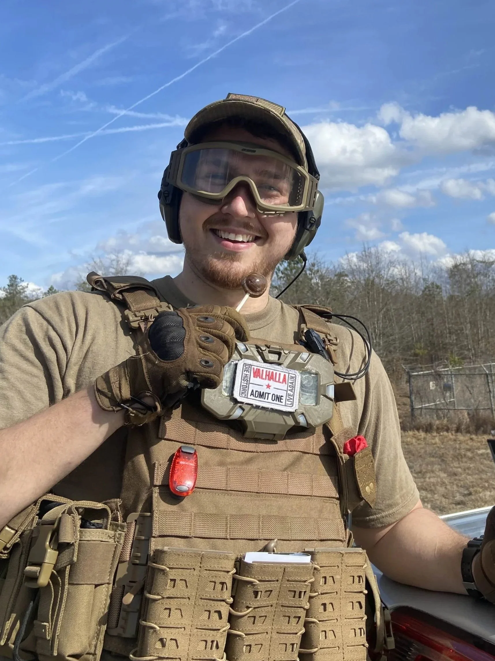 A smiling man in military gear outdoors, wearing tactical vest and goggles, holding a lollipop, with a background of blue sky and trees.