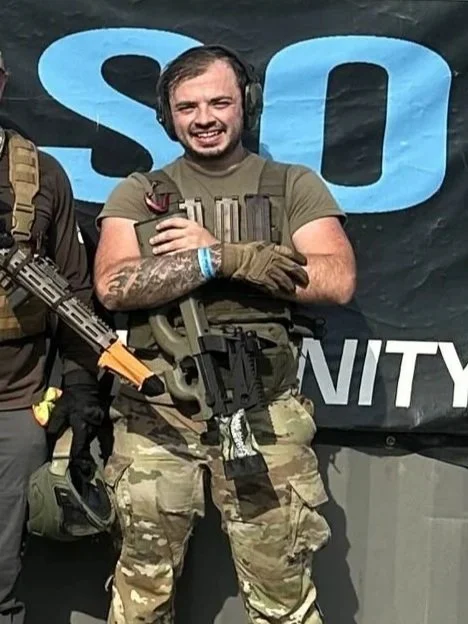 A smiling man in military gear, wearing headphones, camouflage pants, and gloves, holding a rifle, standing in front of a large black banner with blue and white text.
