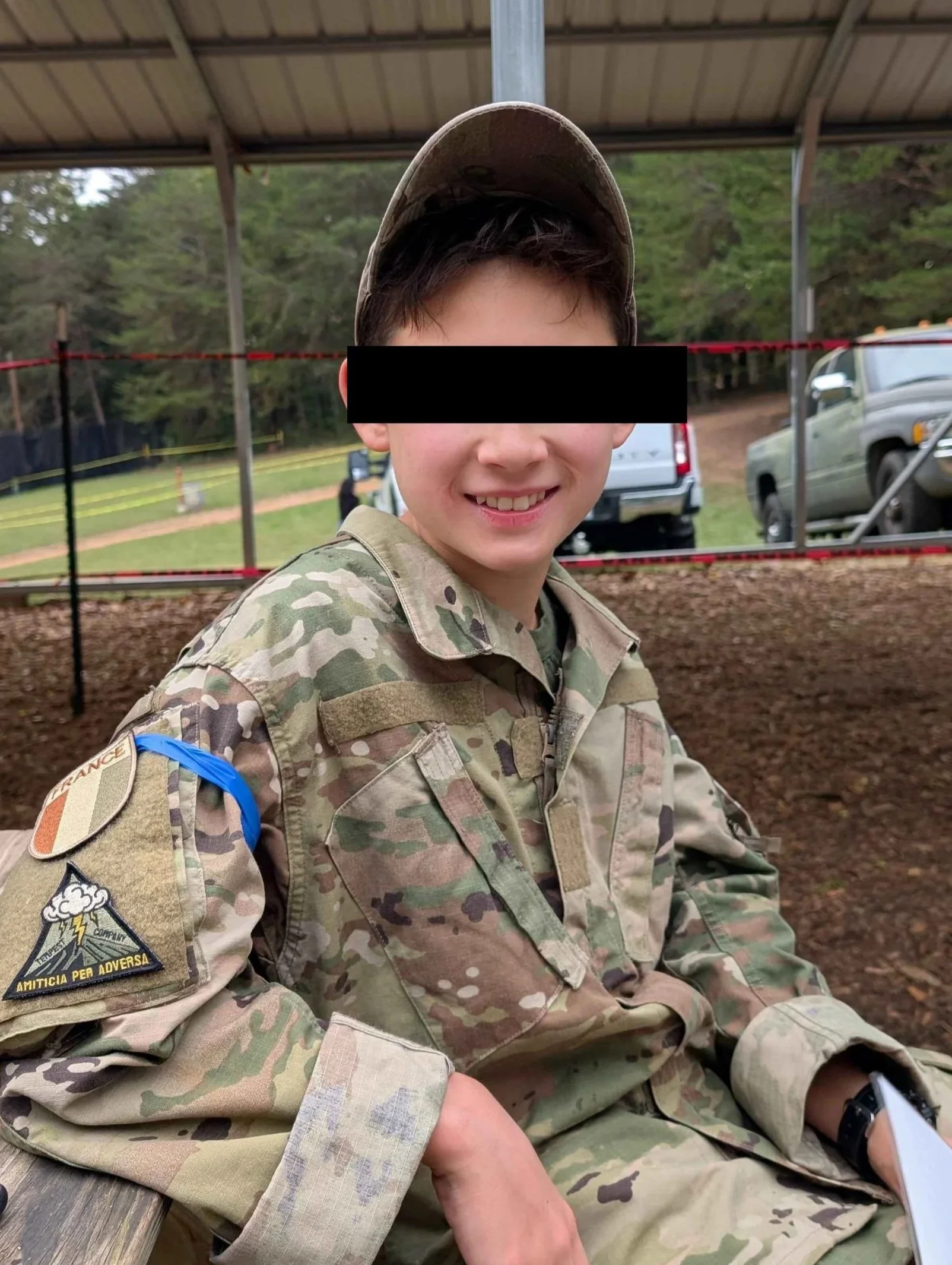 Airsoft player wearing camouflage military uniform and cap, sitting with a smile, blacked out eyes, in an outdoor setting with trees and vehicles in the background.