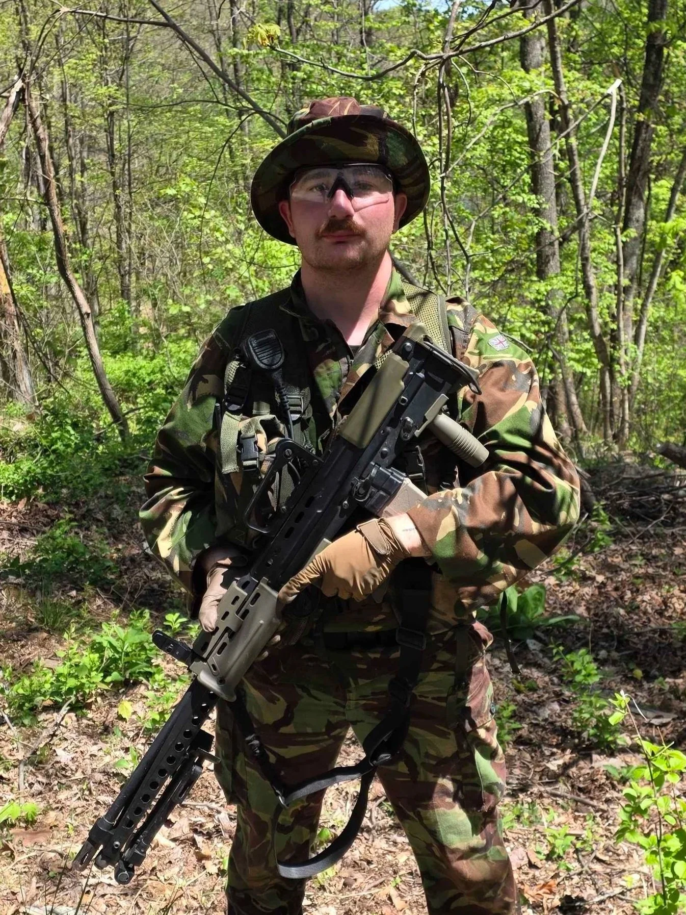 A man in military camouflage uniform, holding a firearm, standing in a forested area with green leaves and trees in the background. He is wearing a camouflage hat and protective glasses.