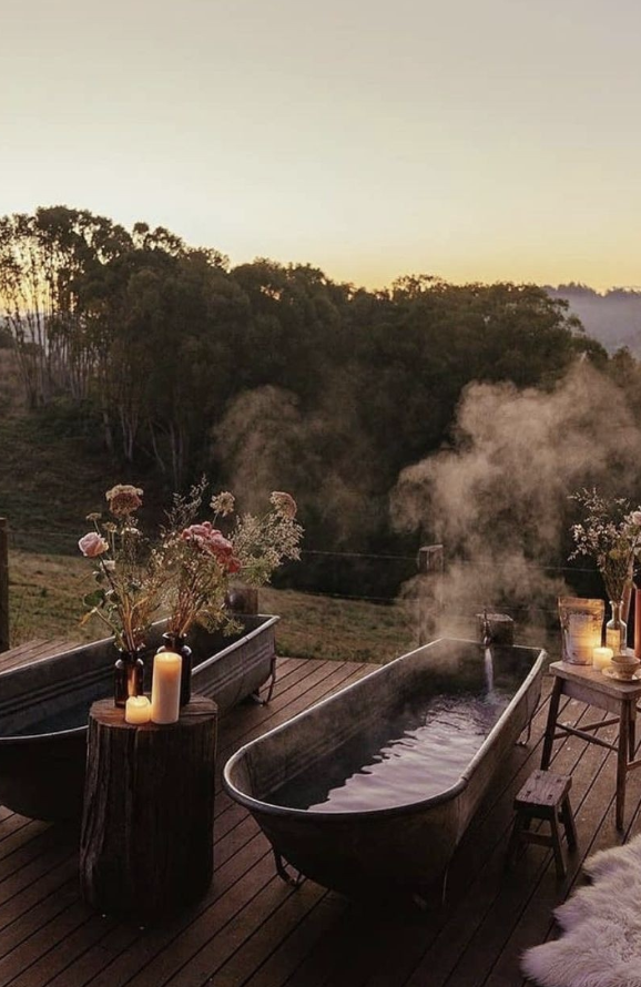 Outdoor wooden deck with two vintage-style bathtubs filled with steaming water, surrounded by candles and flower arrangements, overlooking a misty green forest at sunset.