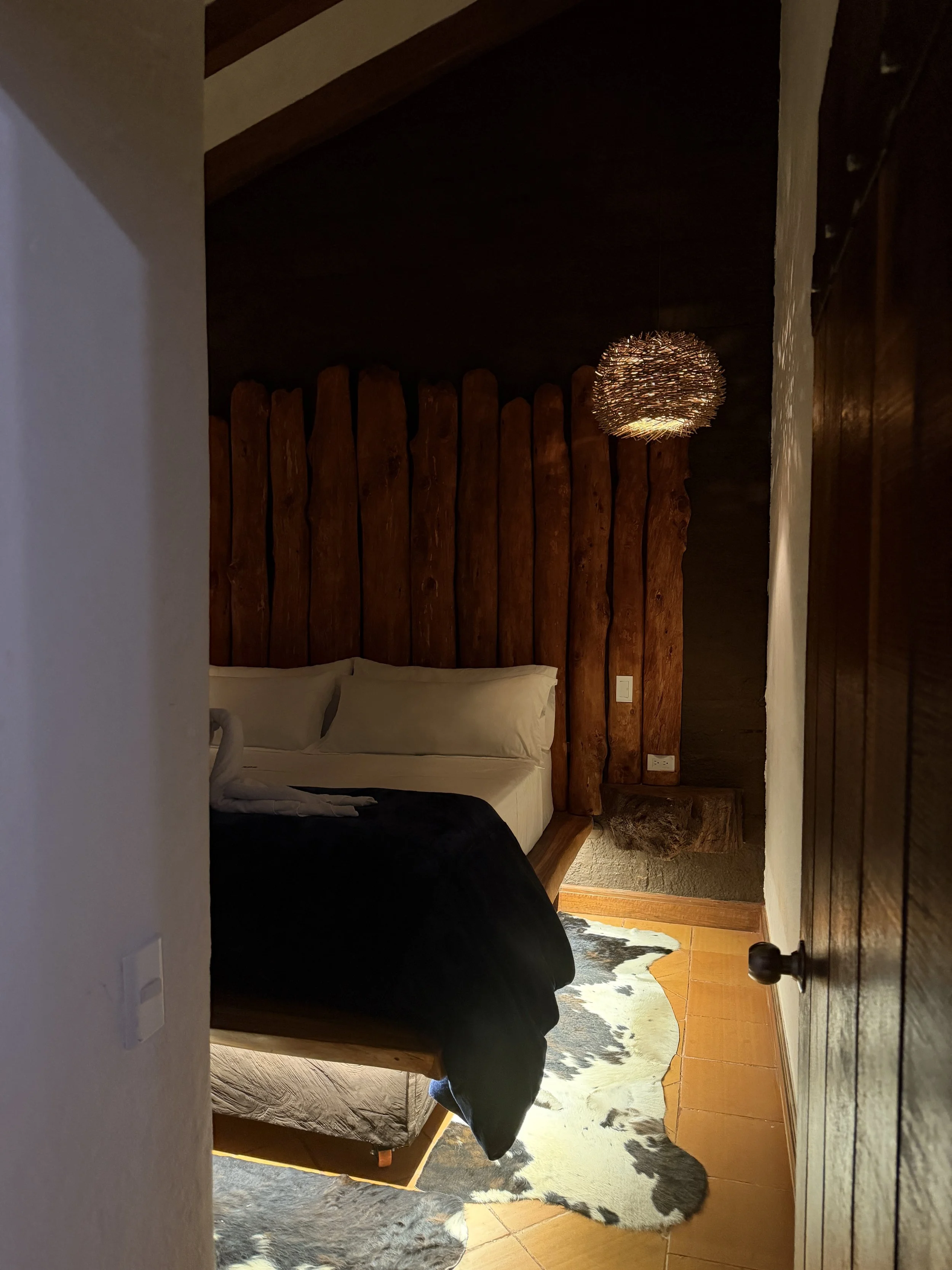 A bedroom with a rustic wooden headboard, a ceiling lamp, a cowhide rug, and a bed with white pillows and a black blanket. The room has warm lighting and wooden flooring.