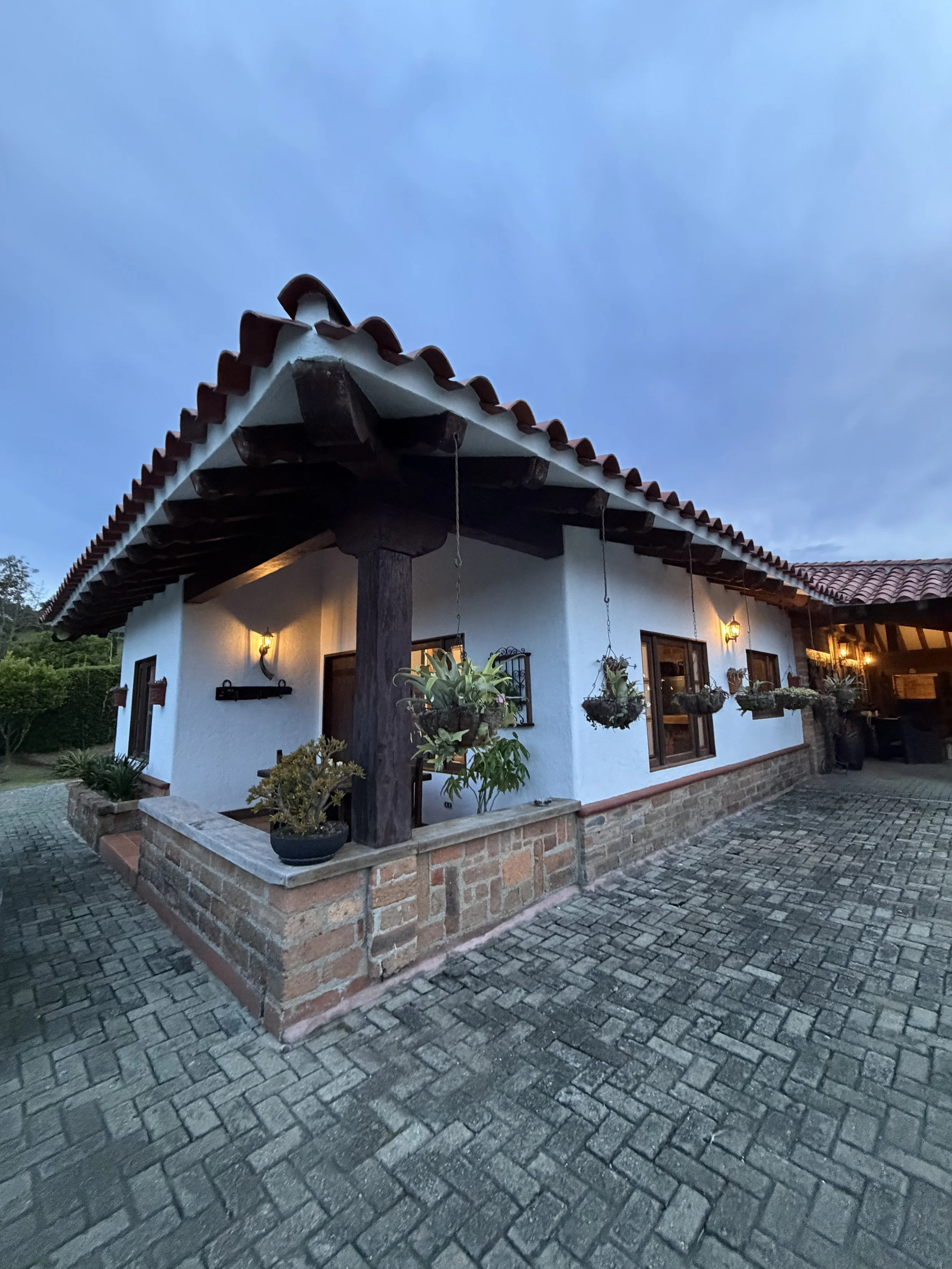Exterior of a house with a clay tile roof, white stucco walls, and hanging plants, illuminated by warm lighting at dusk, with a cobblestone driveway.