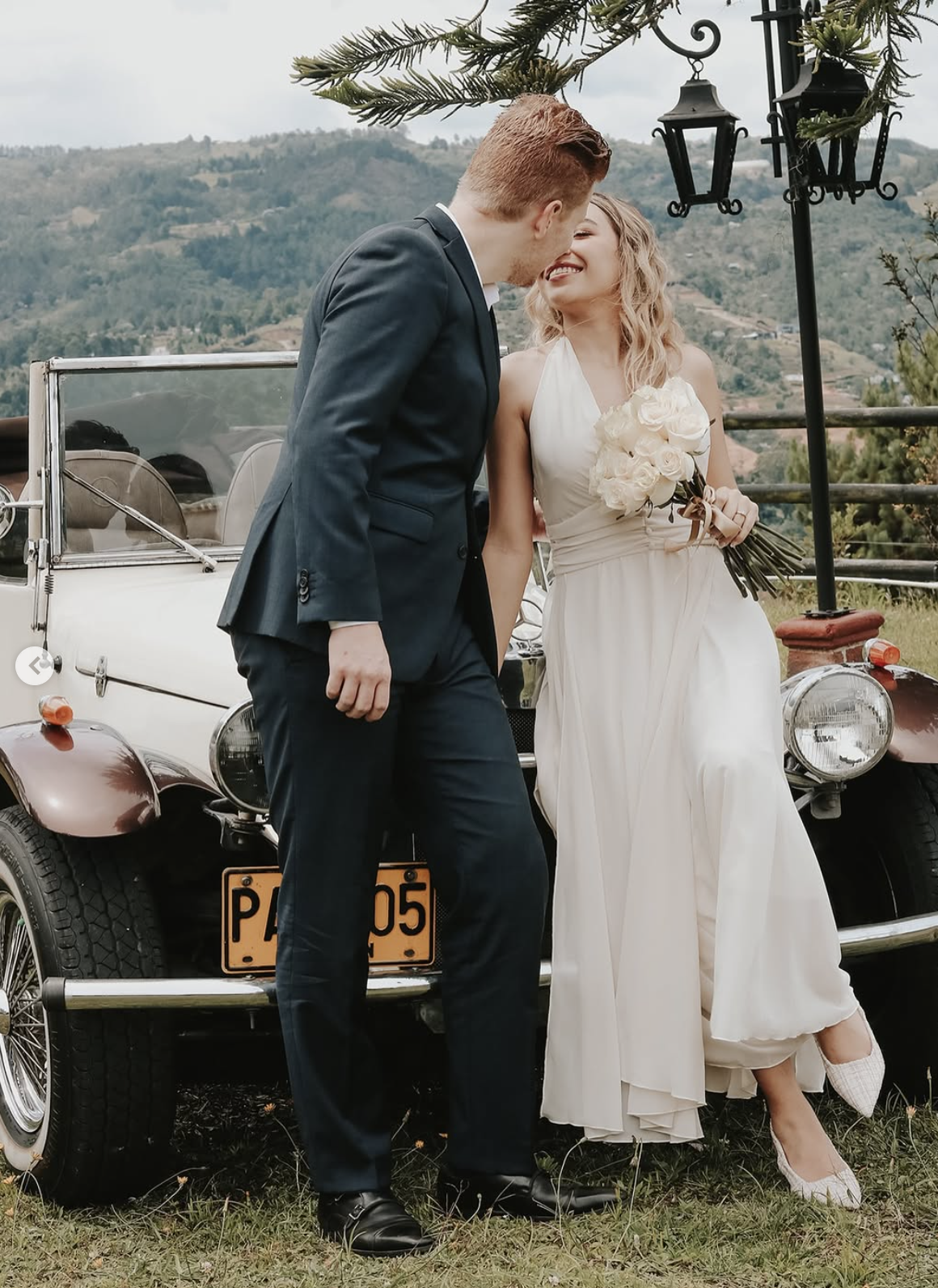 A bride and groom share a tender moment outdoors, standing next to a vintage car with a scenic mountainous background. The bride holds a bouquet of white roses and wears a white dress, while the groom is in a dark suit.