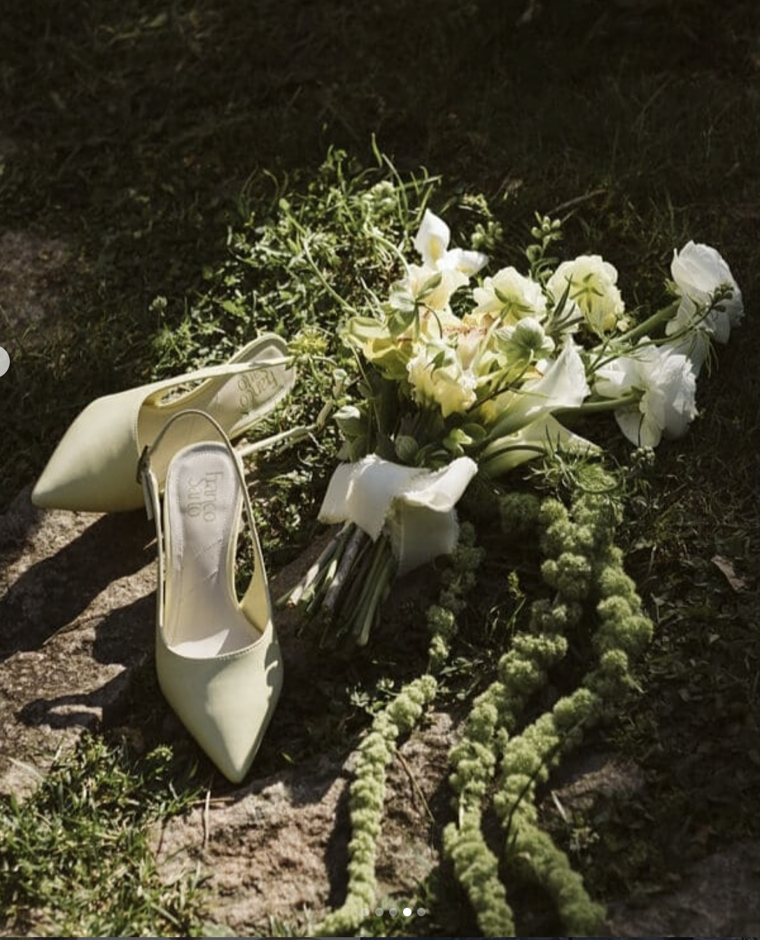A fallen yellow high heel shoe and a bouquet of white flowers on the ground, with greenery and rocky terrain.