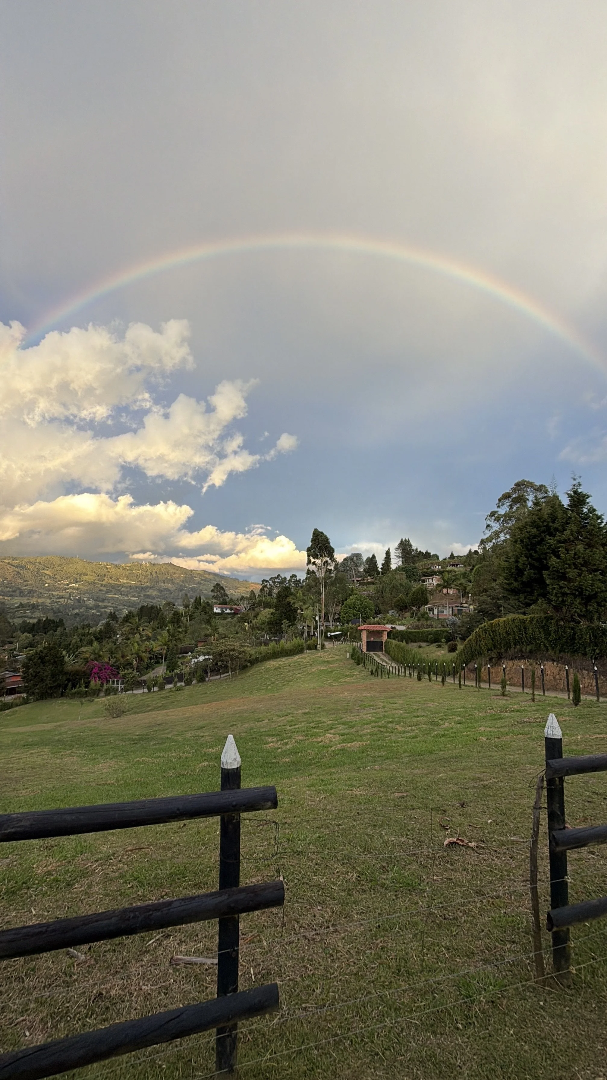 A scenic landscape with a rainbow arching over green hills, houses, trees, and a cloudy sky.