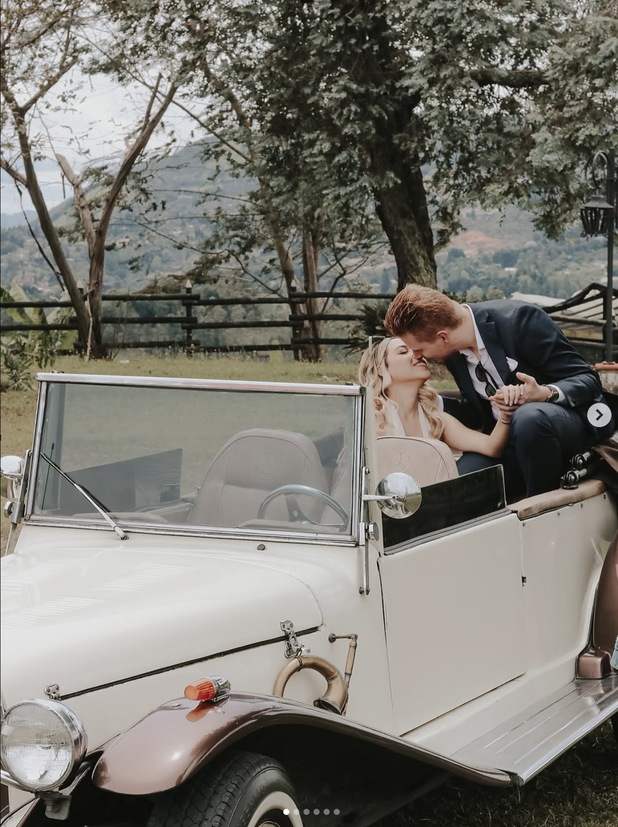 A couple dressed in formal attire sharing an intimate moment in a vintage white convertible car outdoors, surrounded by trees and scenic background.