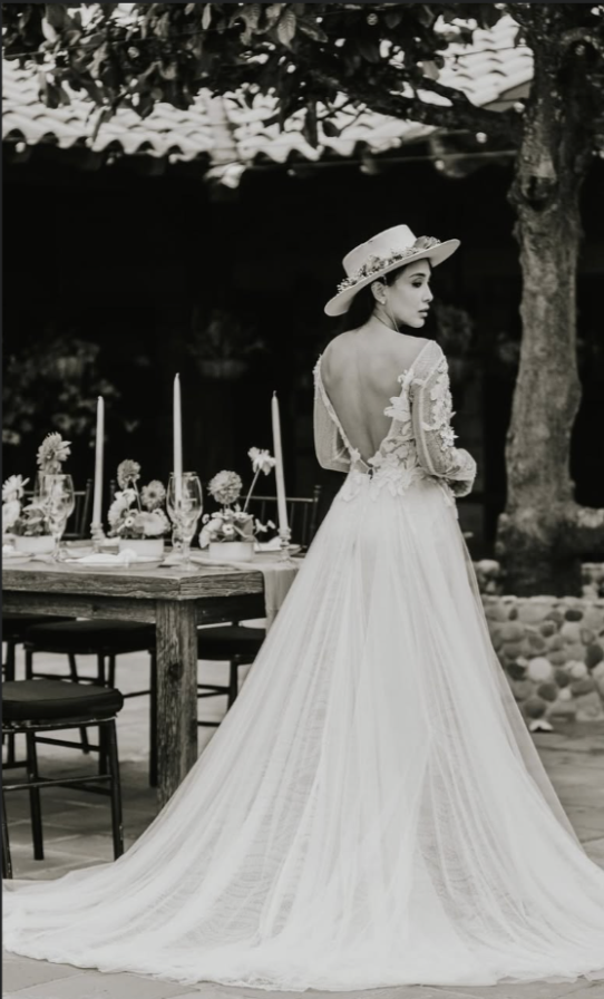 A woman in a wedding dress and wide-brimmed hat standing outdoors near a table with candles and floral arrangements.