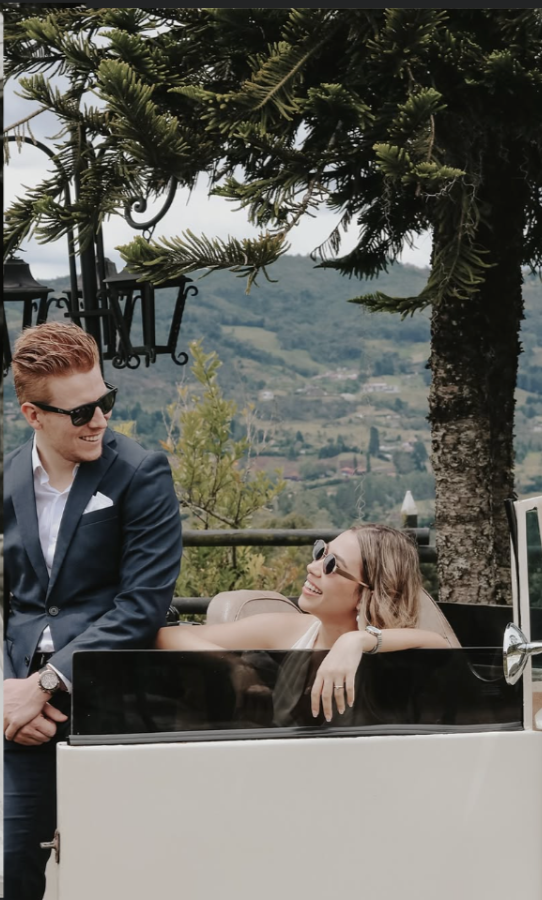 A man in a suit and sunglasses standing next to a woman in sunglasses sitting in an open-top vehicle, with a scenic landscape of trees and hills in the background.