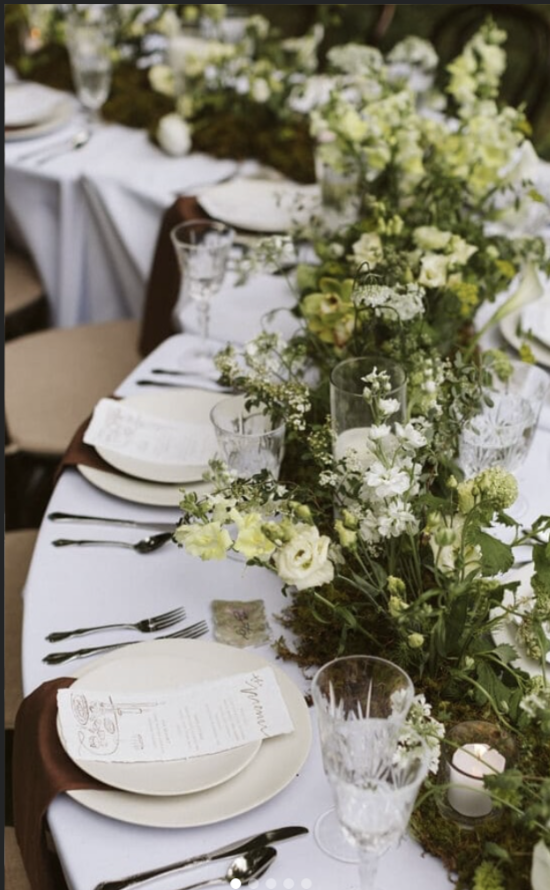 Elegant dining table decorated with white tablecloth and a lush centerpiece of white and green flowers, including roses and other blooms, with glassware, plates, silverware, and a menu card at each setting.