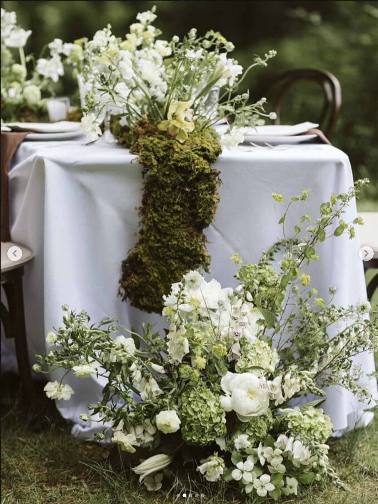 A table decorated with white flowers, greenery, and moss, set outdoors with a white tablecloth and wooden chairs.