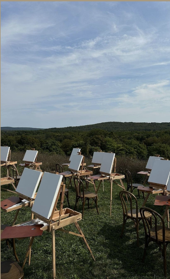 Outdoor painting setup with easels and chairs on grassy field, overlooking rolling hills and blue sky.