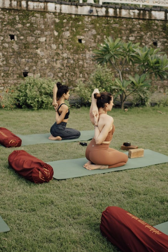 Two women practice yoga outdoors on green mats, performing a seated spine stretch, with trees and a brick wall in the background.