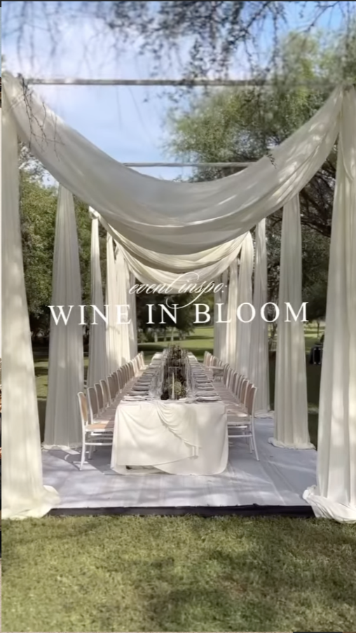 Outdoor wedding reception setup with long dining table, white chairs, and flowing white fabric draped over an open canopy in a park-like setting.