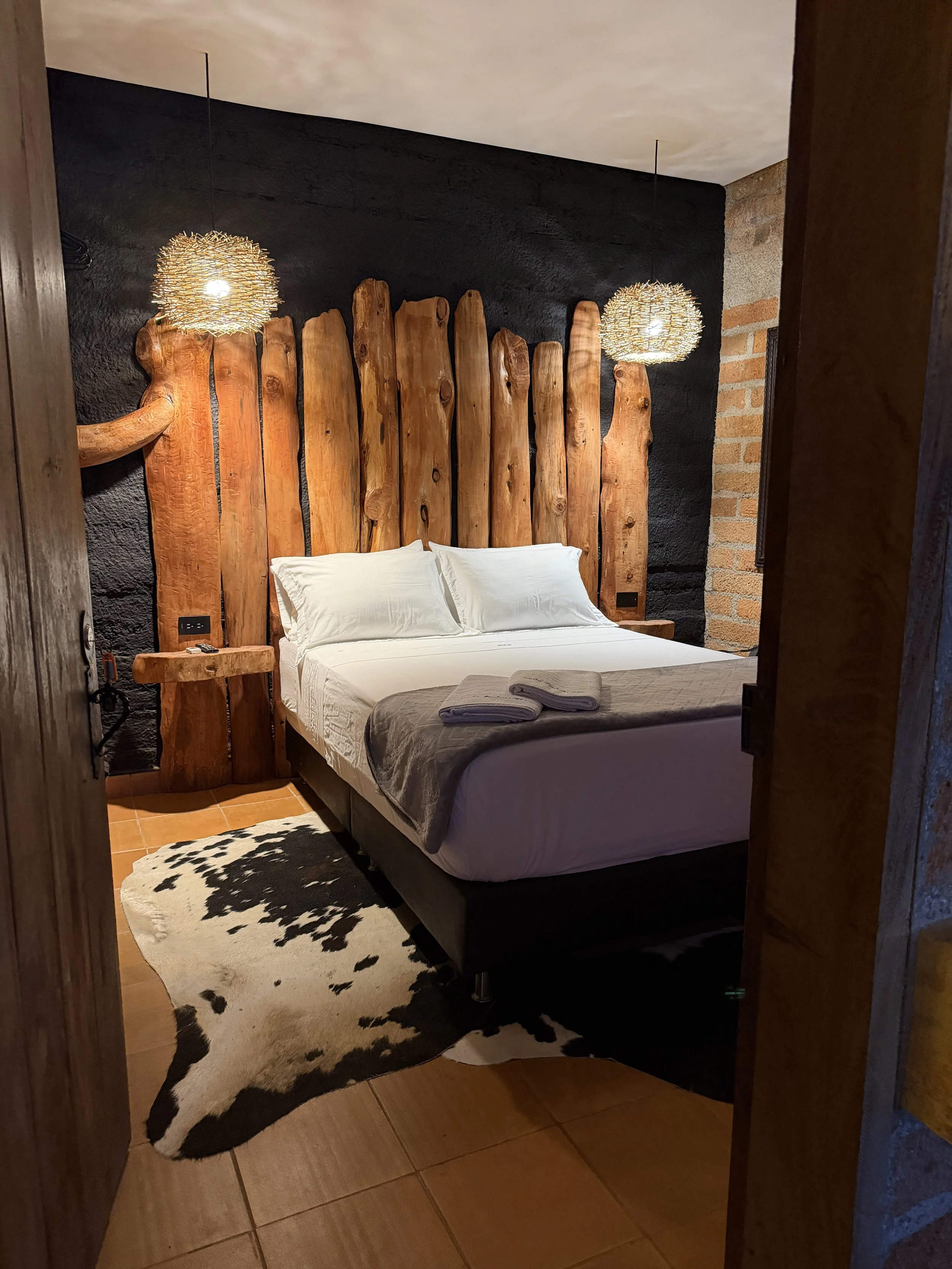 A cozy bedroom with a wooden headboard, white bedding, and a cowhide rug on a tiled floor, illuminated by two woven pendant lights hanging from a dark ceiling.